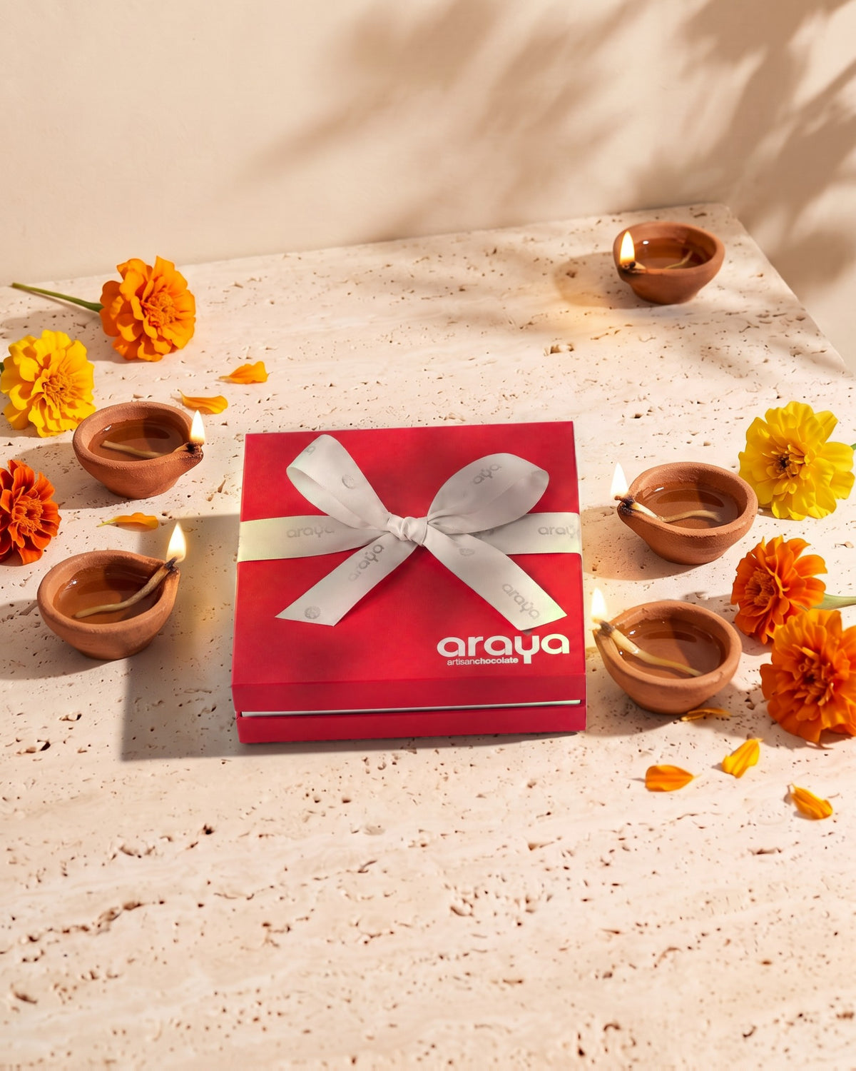 Araya red Diwali gift box with a white ribbon, among lit diyas and marigolds
